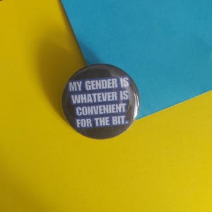 May include: A circular badge with the text "MY GENDER IS WHATEVER IS CONVENIENT FOR THE BIT" in white on a dark brown background. The badge is set against a yellow, blue, and teal background.