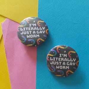 May include: Two circular black badges with the text "I'M LITERALLY JUST A GAY WORM" in white. The badges have a rainbow worm design. The background is a mix of yellow, pink, and blue.