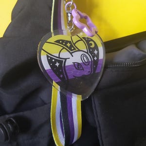 May include: A heart-shaped keyring with a cartoon character design incorporating the non-binary pride flag. It has a purple flower charm and a striped ribbon in yellow, white, and purple. The background is black and yellow.