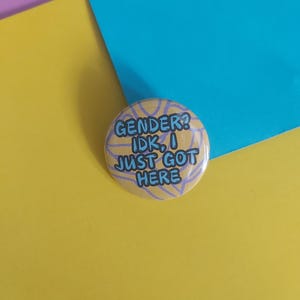 May include: A round button with the text "GENDER? IDK, I JUST GOT HERE" in blue lettering. The button has a yellow background with a light purple pattern. The background is a mix of yellow, blue, and purple.