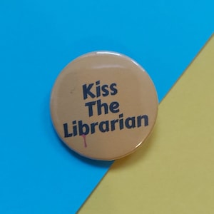 Puede incluir: Un botón amarillo con el texto "Kiss The Librarian" en letras negras. Una pequeña gota roja está debajo del texto.