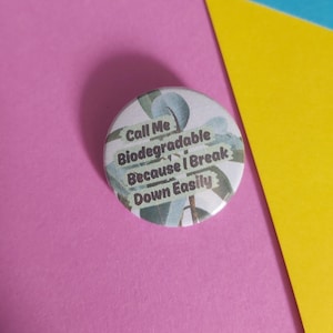May include: A circular badge with the text "Call Me Biodegradable Because I Break Down Easily". The badge has a light green and white floral design. The background is pink, yellow, and blue.
