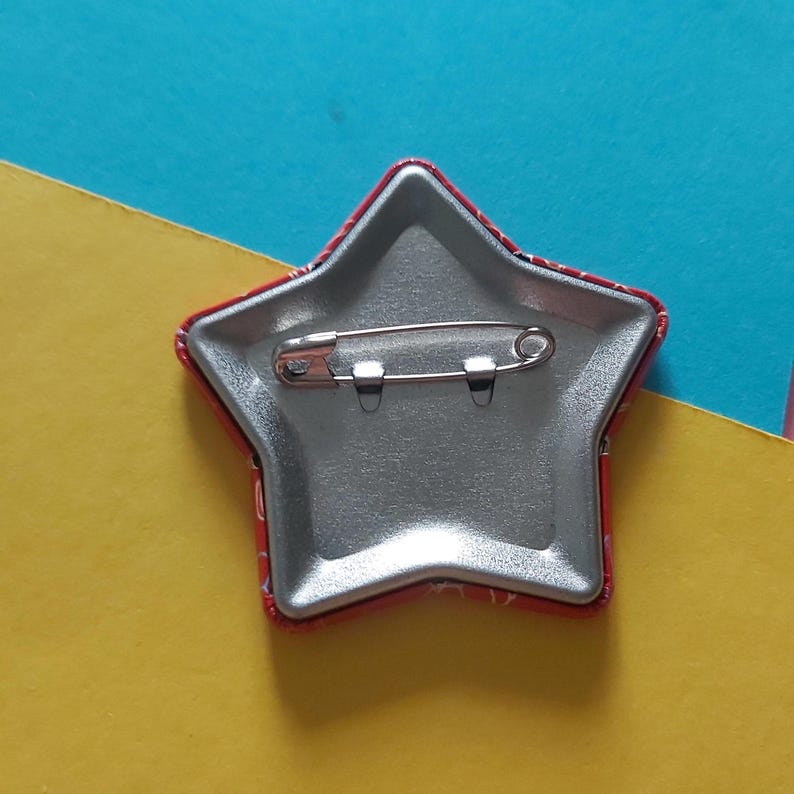 May include: A star-shaped pinback button with a red and white patterned border. The back of the button is silver with a safety pin clasp. The button is on a yellow and blue background.