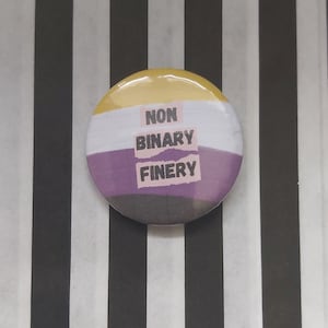 May include: A round button with the non-binary pride flag colors. The button has the words "NON BINARY FINERY" printed on it.