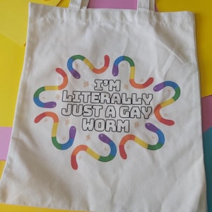 May include: A white tote bag featuring the text "I'M LITERALLY JUST A GAY WORM" in black. The design includes rainbow-coloured worms. The bag has two white handles.