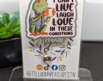 May include: A badge reel with a cartoon frog holding a coffee cup, wearing a vest and shirt. The text on the badge reel says "I CAN'T LIVE LAUGH LOVE IN THESE CONDITIONS". The badge reel is displayed on a clear acrylic stand.