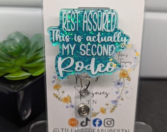 May include: A teal glitter badge reel with the text "REST ASSURED This is actually MY SECOND Rodeo" in white. The badge reel is displayed on a white card with floral accents and the shop's social media handles.