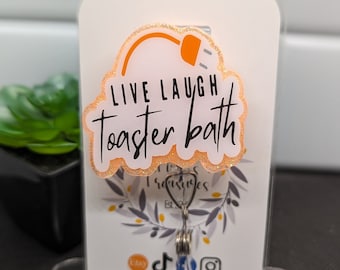 May include: A white card with a peach-colored glitter background and a black outline. The card has a black plug graphic and the text "Live Laugh Toaster Bath". The card is attached to a silver spring clip.