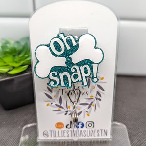 May include: White bone-shaped charm with teal glitter and the text "Oh Snap!" attached to a silver swivel clip. The charm is displayed on a white background with a small plant in the background.