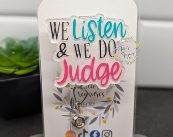 May include: A white acrylic sign with the text "We Listen & We Do Judge" in blue and pink lettering. The sign is on a clear acrylic stand. The sign also includes the text "Tillie's Treasures Est 2021" and social media icons.