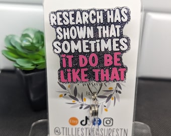 May include: A black and white glitter keychain with the text "Research has shown that sometimes it do be like that". The text is in pink glitter. The keychain is on a white display stand with a floral design and the text "@tilliestreasuresTN".
