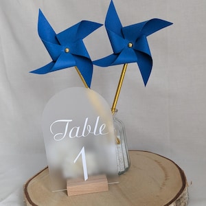 Op de afbeelding: Een tafelnummerdisplay met een mat acrylboog, het woord "Table" en het nummer "1" in witte letters. Twee blauwe windmolens op gouden stelen staan in een glazen vaas. De display staat op een houten schijf.