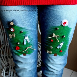 May include: Light blue denim jeans featuring two hand-painted green Christmas trees on the thighs, decorated with small buttons, fabric scraps, and twine bows. The text "by koko4ka JEANS_TUNING_DESIGN" is printed on the upper left thigh area.