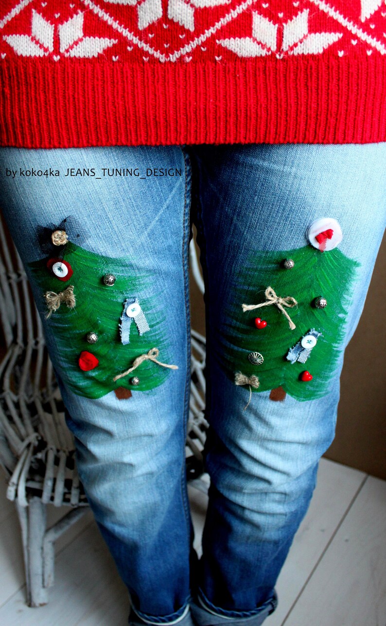 May include: Light blue denim jeans featuring two hand-painted green Christmas trees on the thighs, decorated with small buttons, fabric scraps, and twine bows. The text "by koko4ka JEANS_TUNING_DESIGN" is printed on the upper left thigh area.