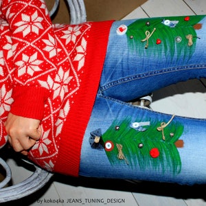 May include: Blue denim jeans featuring hand-painted green Christmas trees on the thighs, decorated with small buttons, fabric scraps, and twine bows. The jeans are paired with a red and white knit sweater with a repeating snowflake pattern.