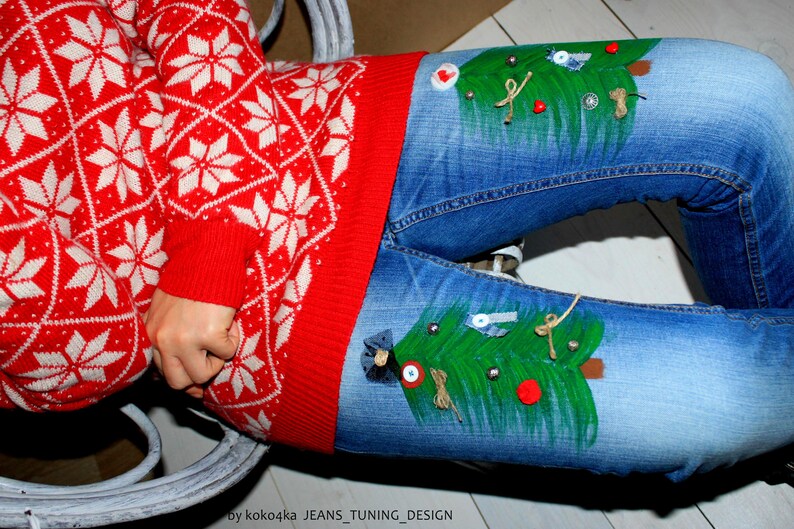 May include: Blue denim jeans featuring hand-painted green Christmas trees on the thighs, decorated with small buttons, fabric scraps, and twine bows. The jeans are paired with a red and white knit sweater with a repeating snowflake pattern.