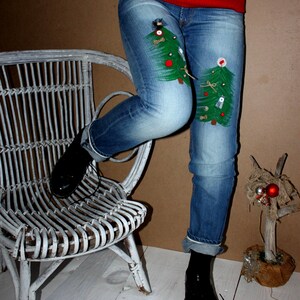 May include: Blue denim jeans featuring two hand-painted green Christmas tree designs on the thighs, decorated with small buttons, beads, and fabric scraps. The jeans are paired with a red and white snowflake patterned sweater and black shoes.