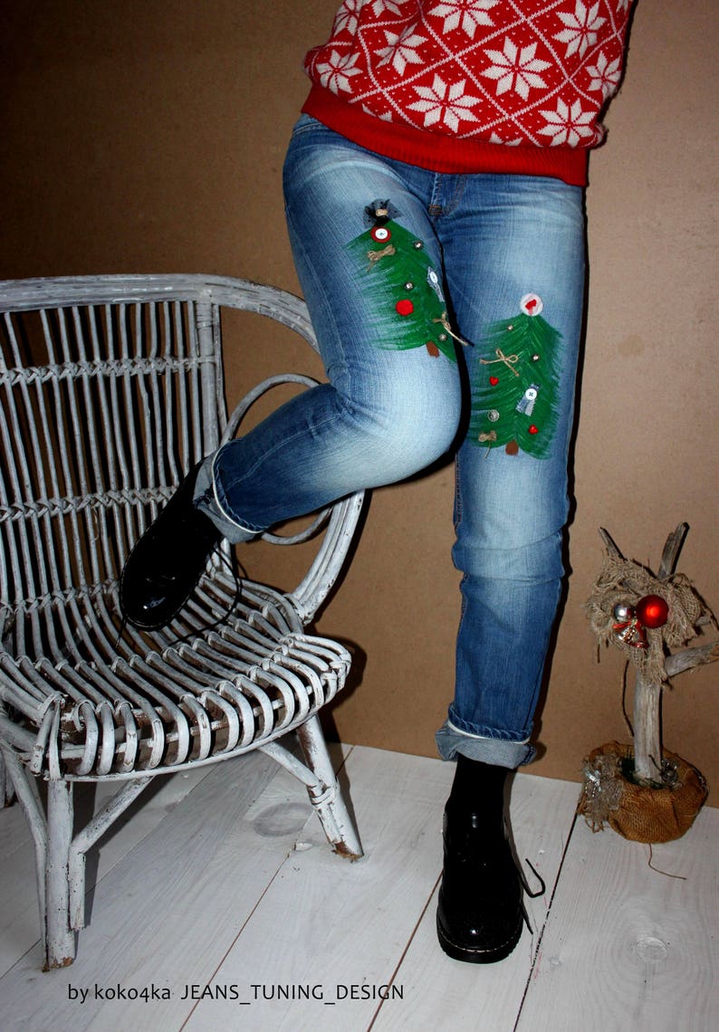 May include: Blue denim jeans featuring two hand-painted green Christmas tree designs on the thighs, decorated with small buttons, beads, and fabric scraps. The jeans are paired with a red and white snowflake patterned sweater and black shoes.