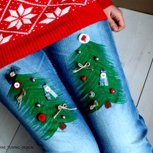 May include: Blue denim jeans featuring two hand-painted green Christmas trees on the thighs, decorated with small buttons, twine bows, and red heart-shaped embellishments. A red sweater with a white snowflake pattern is worn above the jeans.