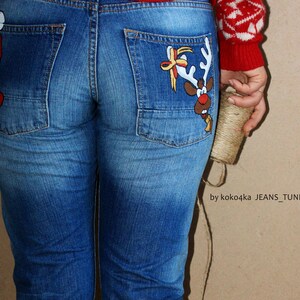 May include: Blue denim jeans featuring hand-painted holiday illustrations on the back pockets. The left pocket displays a Santa Claus figure, while the right pocket features a reindeer with a bow. Text reads by koko4ka JEANS_TUNING_DESIGN.