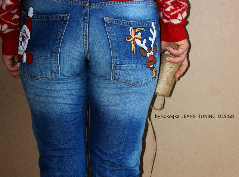 May include: Blue denim jeans featuring hand-painted holiday illustrations on the back pockets. The left pocket displays a Santa Claus figure, while the right pocket features a reindeer with a bow. Text reads by koko4ka JEANS_TUNING_DESIGN.