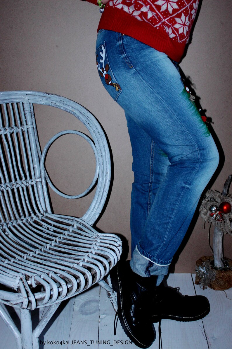 May include: Blue denim jeans featuring a reindeer graphic on the back pocket and a green painted design on the side. The jeans are paired with a red sweater with a white snowflake pattern and black leather boots. Text reads: koko4ka JEANS_TUNING_DESIGN.