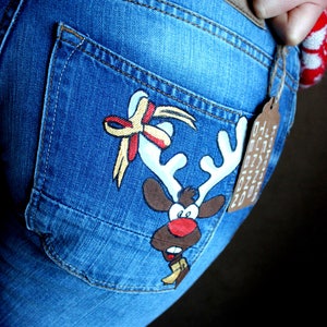 May include: Blue denim jeans featuring a hand-painted cartoon reindeer on the back pocket. The reindeer has white antlers, a red nose, and a yellow scarf. A decorative bow is painted above the antlers. A brown tag reads KOKO4KA JEANS TUNING DESIGN.