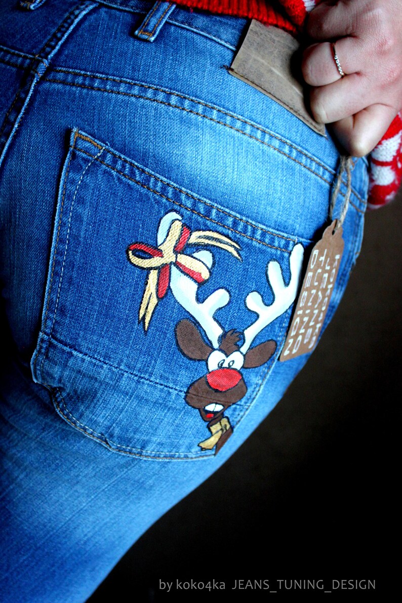 May include: Blue denim jeans featuring a hand-painted cartoon reindeer on the back pocket. The reindeer has white antlers, a red nose, and a yellow scarf. A decorative bow is painted above the antlers. A brown tag reads KOKO4KA JEANS TUNING DESIGN.