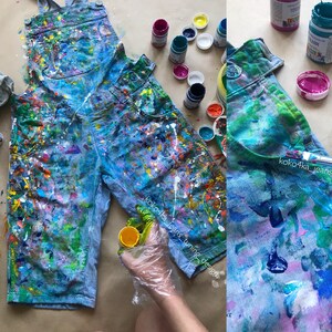 Puede incluir: Mono vaquero azul claro cubierto de salpicaduras de pintura coloridas. La pintura incluye tonos de azul, verde, rosa, amarillo y blanco. Hay botes de pintura y suministros cerca. El texto "koko4ka_jeans_tuning_design" es visible.