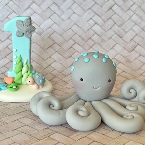 May include: A light blue number one cake topper decorated with a grey flower, green seaweed, a small turtle, and a shell, alongside a grey octopus figurine with light blue spots on its head and curled tentacles, both crafted from smooth material.