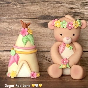 May include: A pink and yellow teepee cake topper with a brown stick on top and green leaves. A brown teddy bear cake topper with a pink and yellow flower crown and a pink feather.