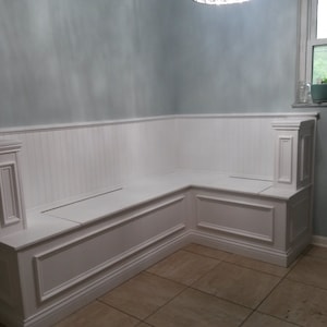 Peut inclure: Banc intégré d'angle blanc avec côtés et dossier à panneaux. Le banc présente des moulures décoratives et une assise plate, idéal pour une cuisine ou un coin repas. Le banc est peint en blanc et est placé contre un mur bleu clair.
