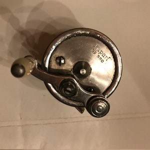 Könnte beinhalten: Eine silberne Angelrolle mit einem weißen Griff. Die Rolle trägt den Text "Tripart No 580".