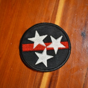 May include: A black and red embroidered patch with three white stars. The stars are arranged in a triangle shape with a red stripe running horizontally through the middle.