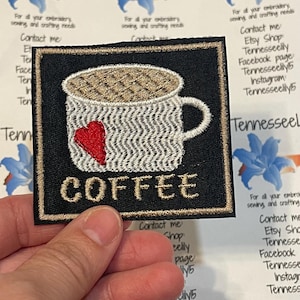 May include: Embroidered patch featuring a coffee cup with a red heart and the word "COFFEE" in gold thread. The patch is black with a gold border. The background includes text and floral designs.
