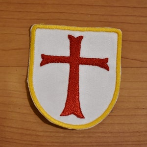 May include: A white fabric patch with a red cross in the center. The patch is surrounded by a yellow border and has a shield shape.