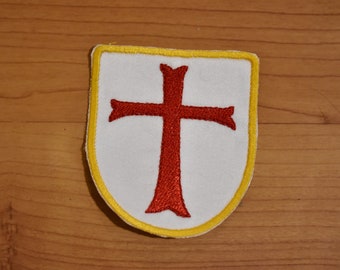 May include: A white fabric patch with a red cross in the center. The patch is surrounded by a yellow border and has a shield shape.