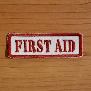 May include: White rectangular patch with a red embroidered border and the text "FIRST AID" in red.