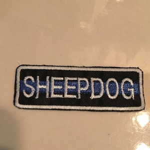 May include: A black and white embroidered patch with the word "SHEEPDOG" in white lettering. A thin blue line runs horizontally across the patch.