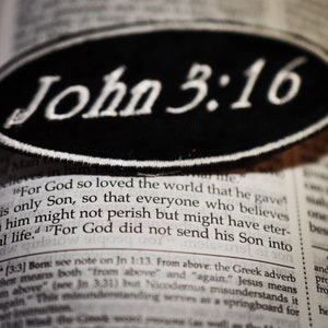 May include: A close-up of a black and white patch with the text "John 3:16" in white lettering. The patch is lying on top of an open book.