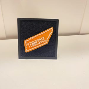 May include: An orange embroidered patch in the shape of the state of Tennessee with a white stitched outline and the word TENNESSEE in white lettering, mounted on a dark blue square display board with a black frame.