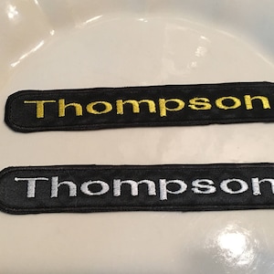 May include: Two black fabric patches with the name "Thompson" embroidered in gold and white thread.