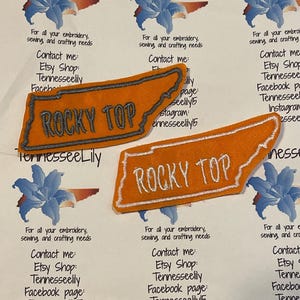 May include: Two orange embroidered patches in the shape of Tennessee, with the words "ROCKY TOP" stitched in white. The patches are surrounded by text and floral graphics, advertising embroidery and crafting services.