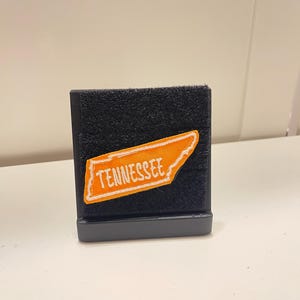 Könnte beinhalten: Ein schwarzer, rechteckiger Präsentationsständer mit einer strukturierten Klettverschluss-Oberfläche. In der Mitte befindet sich ein orangefarbener, gestickter Aufnäher in Form des Bundesstaates Tennessee mit dem weißen Schriftzug TENNESSEE.