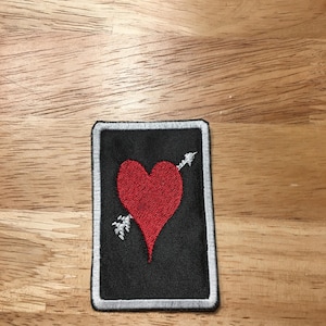 May include: A red heart with an arrow through it is embroidered on a black patch with a white border.