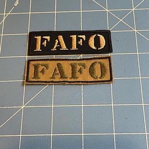 May include: Two rectangular embroidered patches. One is black with gold text reading "FAFO". The other is tan with olive green text reading "FAFO". Both patches are on a blue grid surface.