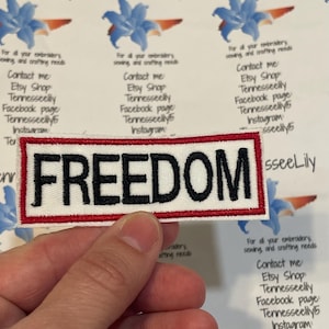 May include: A rectangular patch with the word "FREEDOM" in black capital letters on a white background, framed by a red border. The patch is held by a hand, with other printed materials in the background.