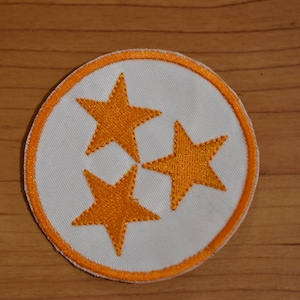 May include: Orange embroidered stars on a white circular patch with an orange border. The stars are arranged in a triangular pattern.