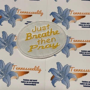 May include: An oval patch with the embroidered phrase "Just Breathe then Pray" in gold thread. The patch is on a white background. The background also features blue flower designs and the text "Tennesseelily".