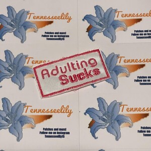 May include: A sheet of stickers featuring a blue lily design and the text "Tennesseelily". One sticker has a patch that says "Adulting Sucks" in red embroidery. The stickers also include the text "Patches and more! Follow me on Instagram Tennesseelily15".